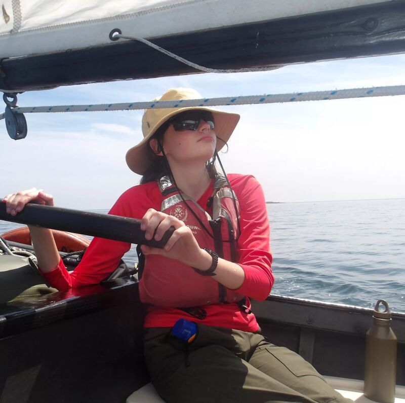 A woman wearing a hat, sunglasses, and a red long-sleeved shirt is sitting in a small boat on the water. She is holding a black paddle with both hands and looking off to the side. She is also wearing a life vest. There is a water bottle next to her. The boat appears to be under a sail.